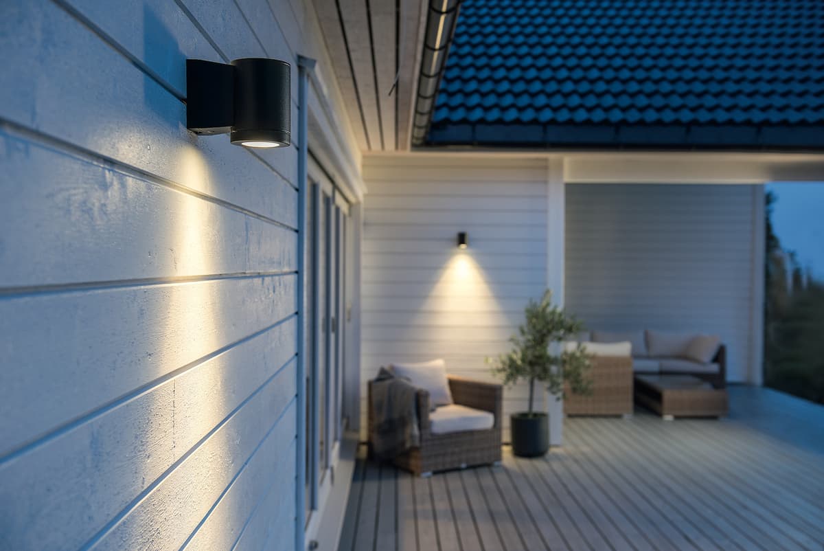 Veggmontert utelys foran en veranda med hagemøbler og en plante Veggmontert utelys foran en veranda med hagemøbler og en plante