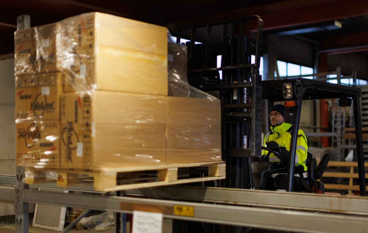 Mann i arbeidsklær løfter paller med pappesker med en gaffeltruck Mann i arbeidsklær løfter paller med pappesker med en gaffeltruck