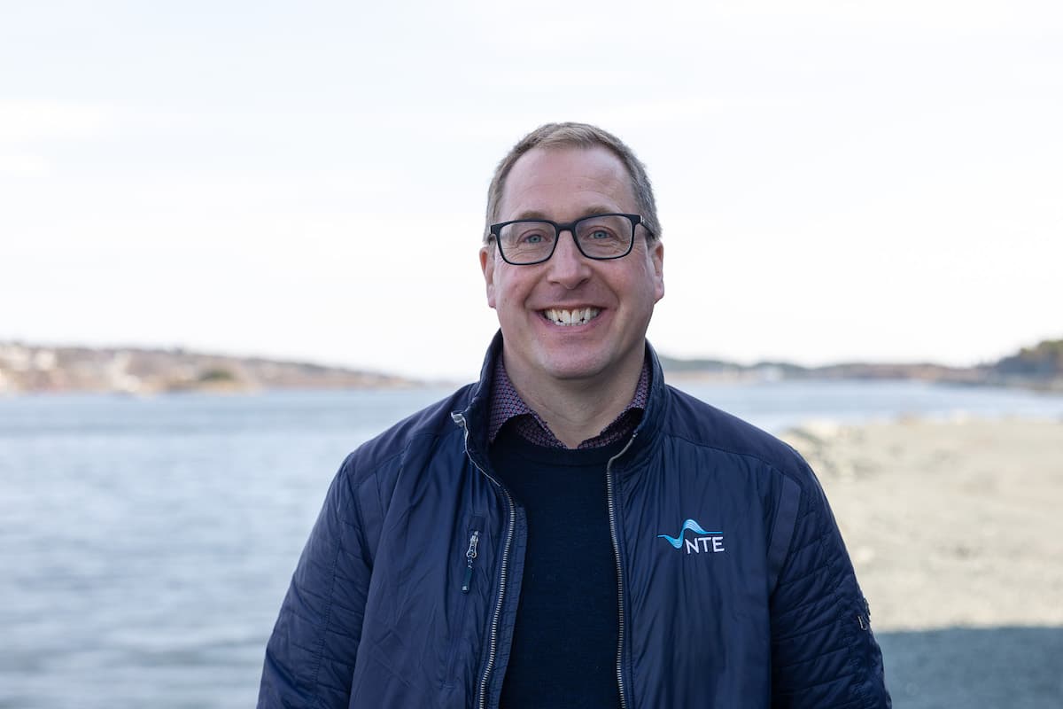 Jakob Hahre Krogsrud NTE Smilende mann med briller og jakke med NTE-logoen på står foran et vann