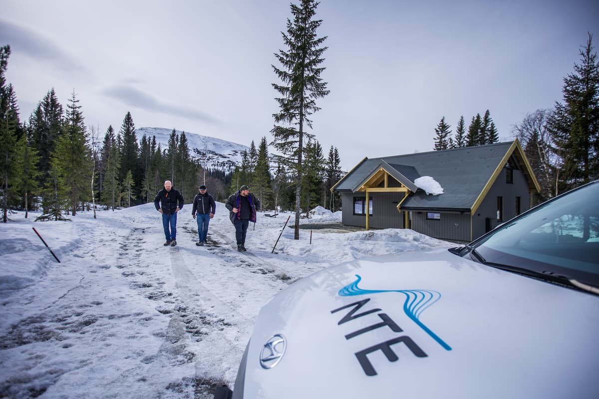 Hytter ved Teveltunet Tre menn går mot en bil med NTE-logoen på panseret. I bakgrunnen en hytte, snø, fjell og skog.