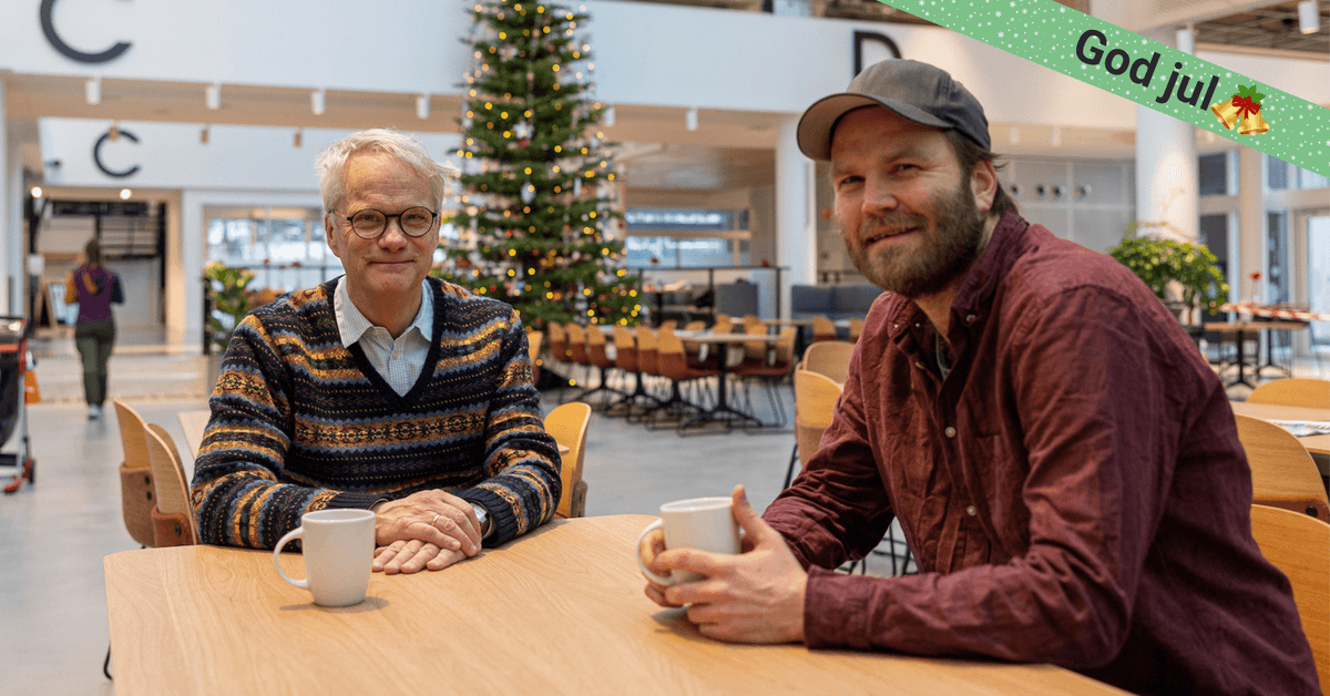forside To menn med hver sin kaffekopp smiler, med et stort juletre i bakgrunnen