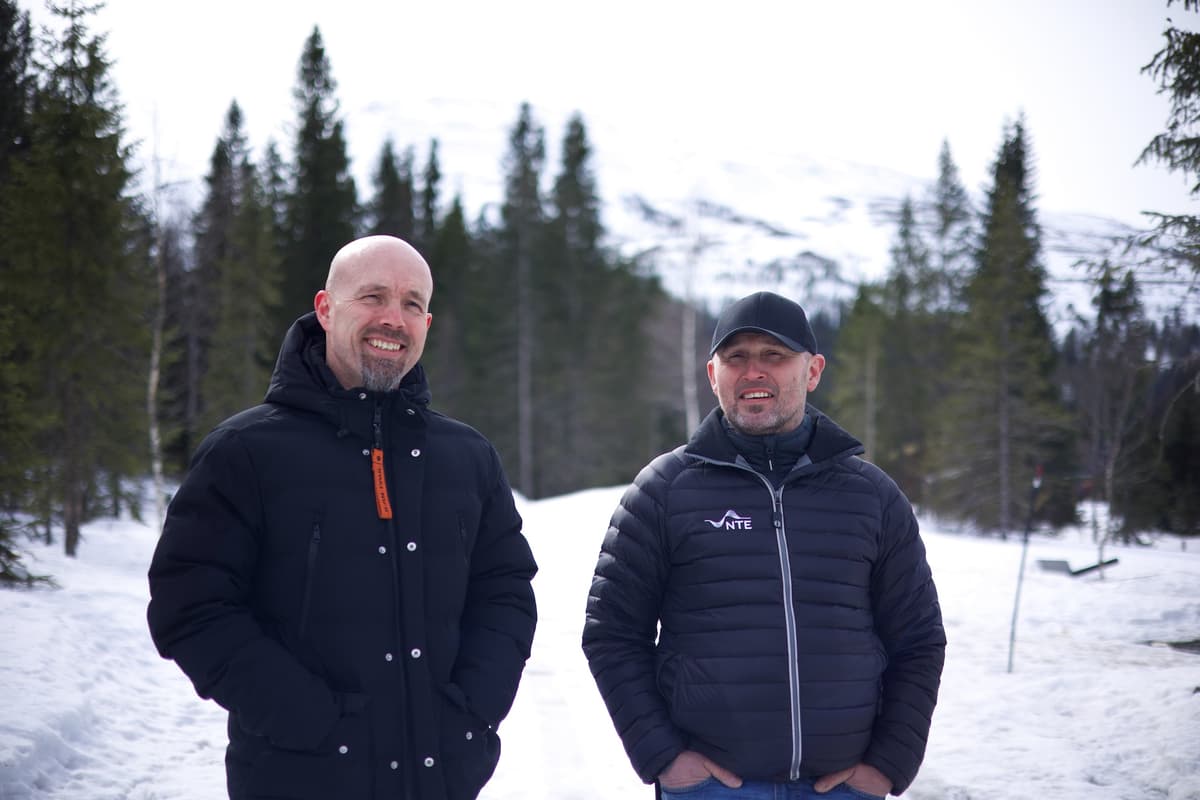 Stian Kilaas og John Ellingen To smilende menn; en med caps, står på en skogsvei i snø med snødekte fjell og grantrær bak seg