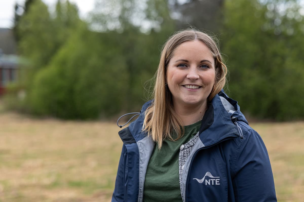 ranja vassli Smilende dame med halvlangt hår og jakke med NTE-logoen på står foran en treklynge