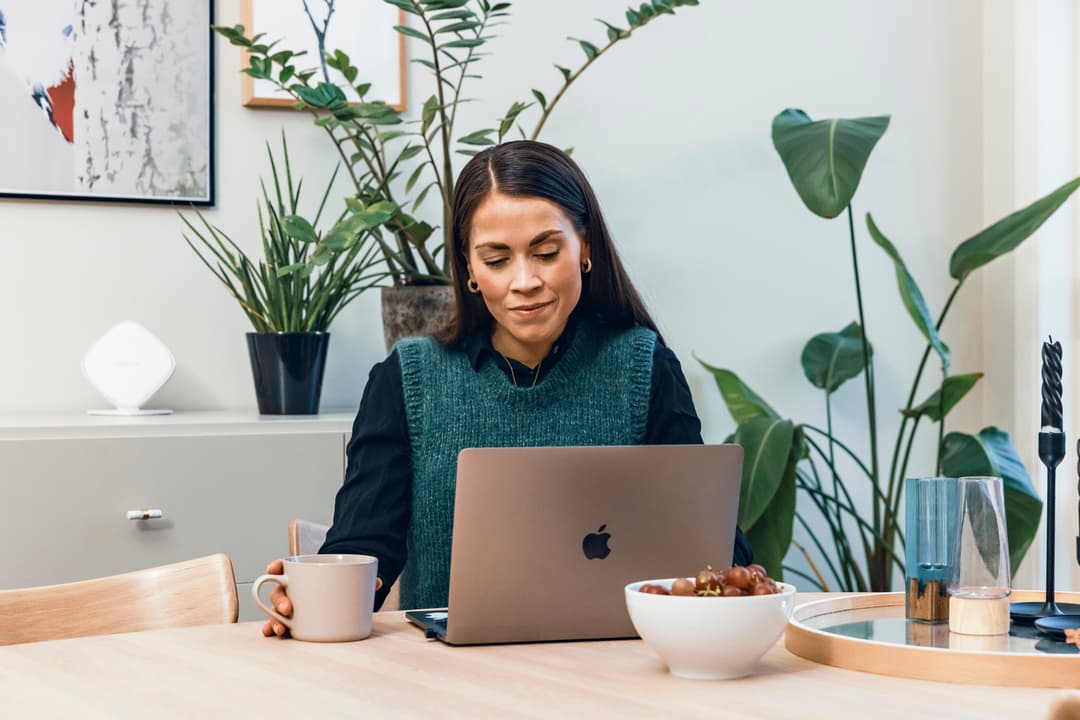 Dame som jobber på hjemmekontor Same som sitter ved et bord. Hun ser på en laptop foran seg, og i hånden holder hun en kaffekopp. Bilde.
