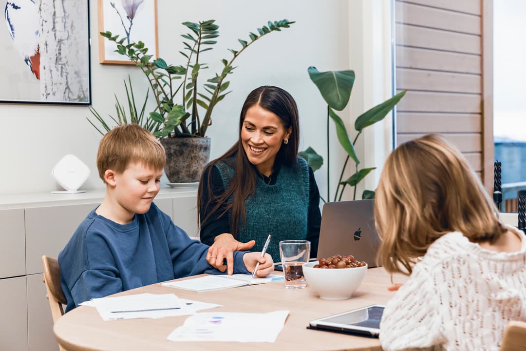 Bilde av mor og to barn som sitter ved et bord. Mor hjelper sønnen med lekser. Datteren ser på et nettbrett. Bilde av mor og to barn som sitter ved et bord. Mor hjelper sønnen med lekser. Datteren ser på et nettbrett.