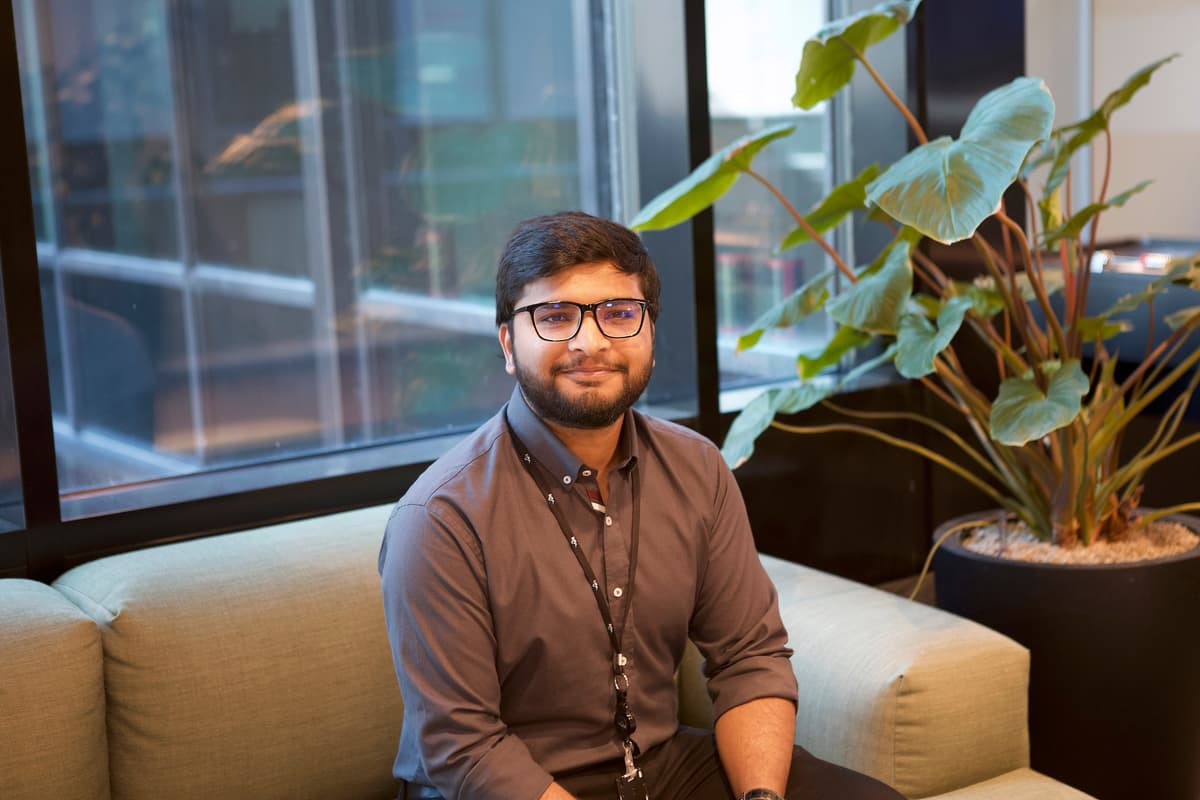 Hamza Javeed Smilende mann med briller sitter i en sofa som står ved siden av en stor grønnplante