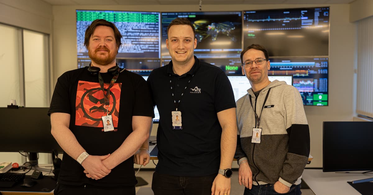 gutta på Nocen Tre smilende menn foran en vegg dekket av skjermer